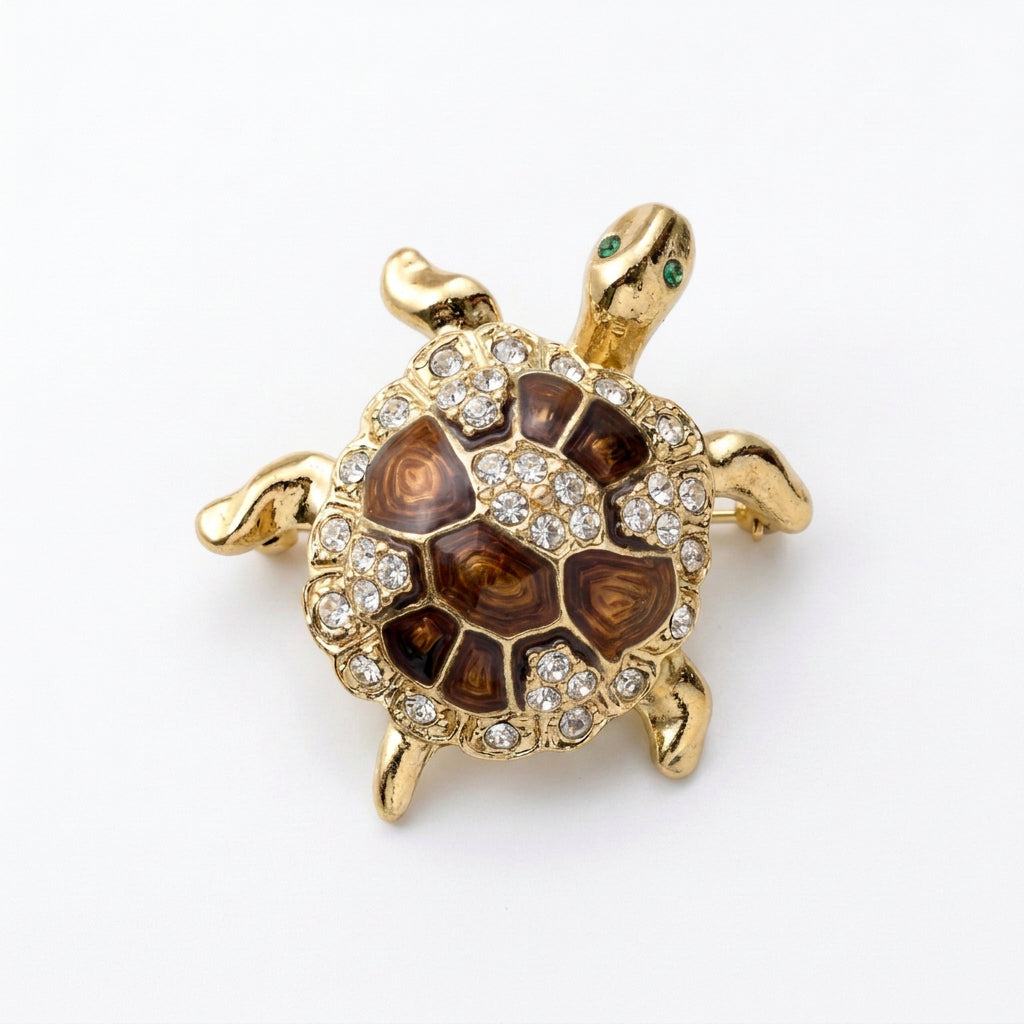 A gold turtle-shaped brooch featuring brown enamel scales, a border of clear rhinestones, and small green crystal  eyes.