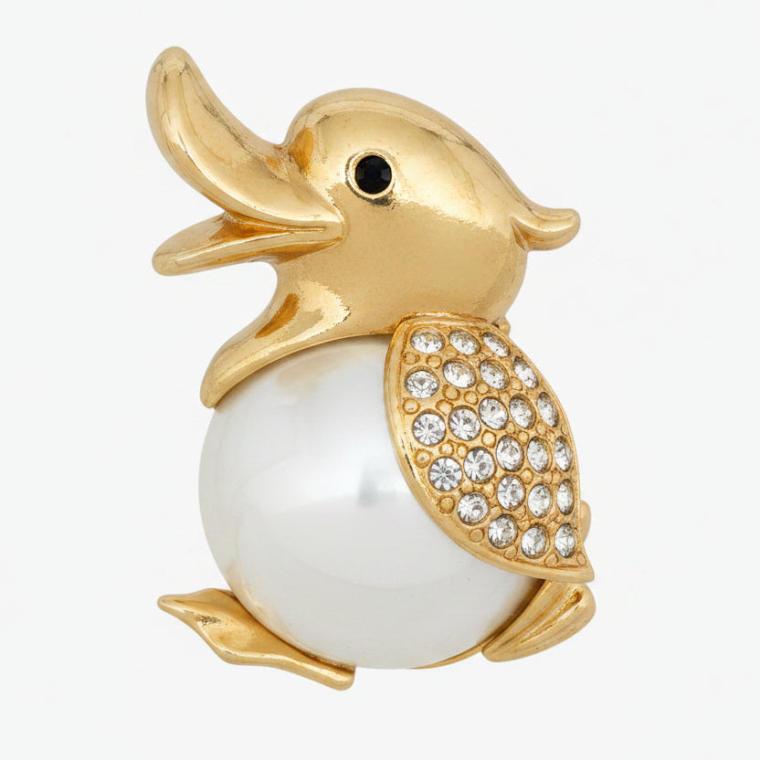 A gold plated duck brooch with a glossy white pearl body and a wing encrusted with clear rhinestones, shown on a white background