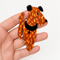 A hand holding a Lea Stein  dog-shaped brooch with an orange and brown spotted pattern, black ears, and a black collar against a plain light background.