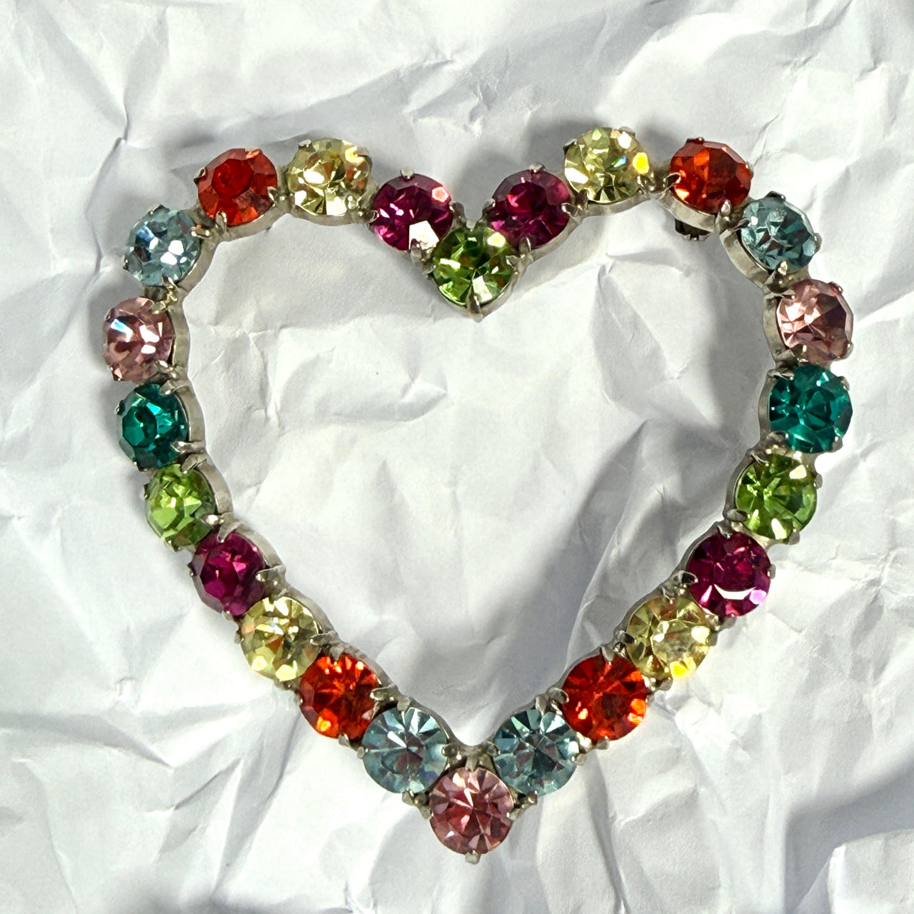 A heart-shaped brooch made of colourful round rhinestones in red, orange, pink, green, blue, and yellow, set in silver-toned metal, displayed on a crumpled white paper background.