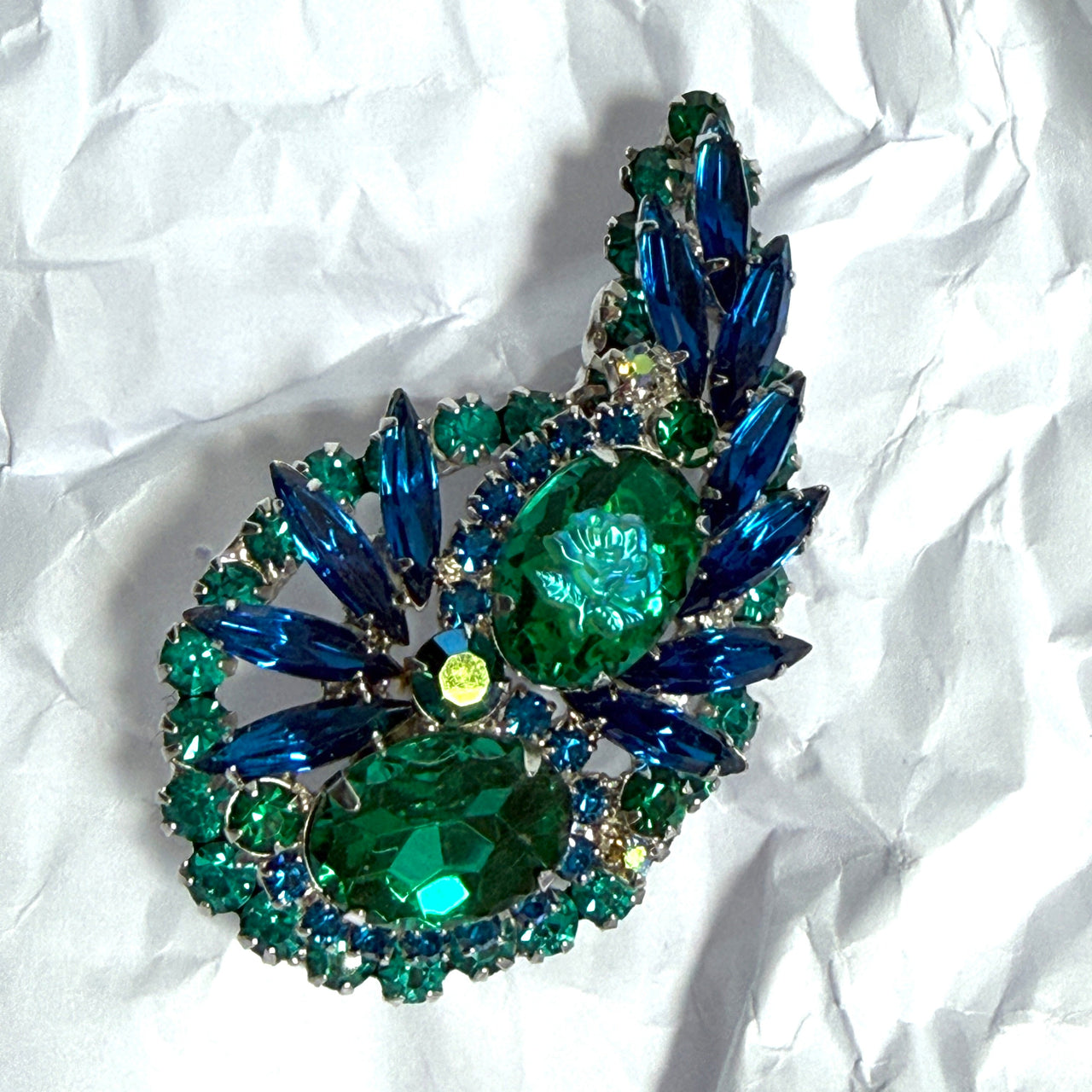A vintage brooch featuring large green and blue faceted rhinestones arranged in a teardrop or leaf-like shape, resting on a crumpled white paper background.