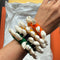 Three colorful woven bracelets adorned with white cowrie shells, worn on a person's wrist over a white tissue and orange surface.