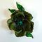 A vintage-style enamel flower brooch featuring dark green and black striped petals with a small green crystal center, set against a plain off-white background.